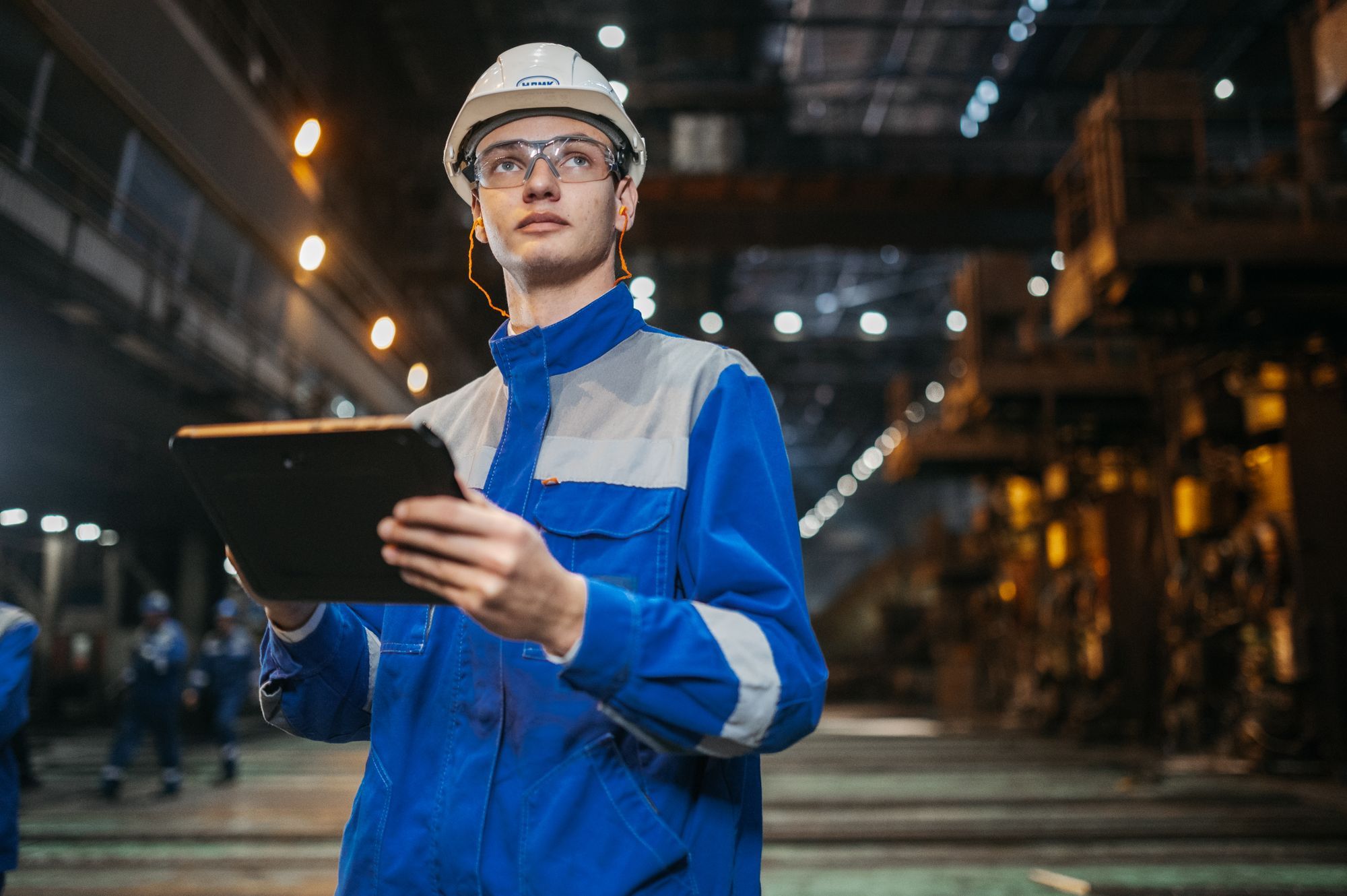 An engineer in safety glasses and hard hat reviewing audit evidence on a tablet in a factory
